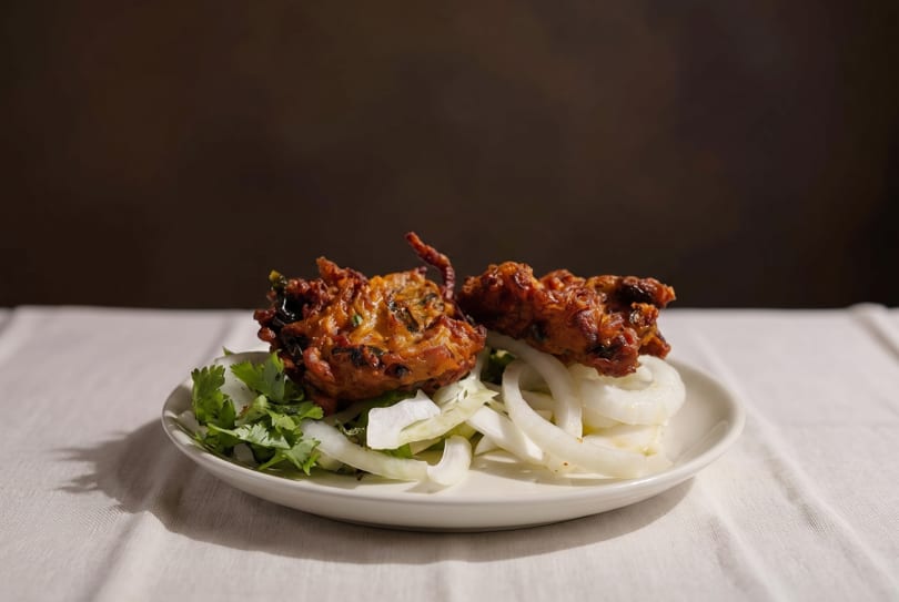 Two golden-brown pakoras, garnished with fresh cilantro and sliced onions, on a white plate.