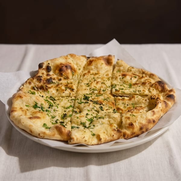A round, golden-brown naan bread, cut into squares and topped with green herbs, on a white plate.