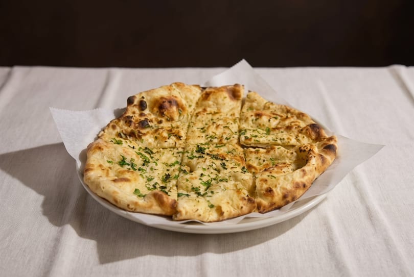 A round, golden-brown naan bread, cut into squares and topped with green herbs, on a white plate.